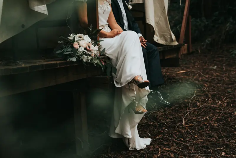 A seated wedding couple holding hands beside a bouquet
